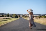 A woman in a long patterned dress and wide-brimmed hat walking down a deserted rural road. The road stretches into the distance, flanked by fields and fences, under a clear blue sky. The surrounding landscape includes grass and shrubs with some rolling hills in the background.