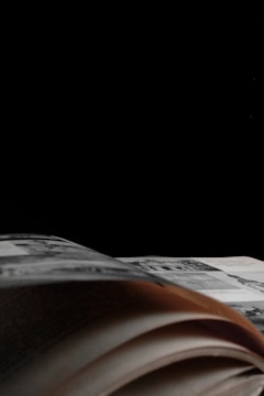 A partially open book with visible printed pages in a low-light setting. The background is predominantly black, creating a dramatic contrast with the pages.