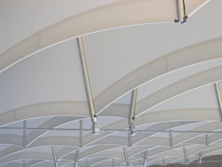 Curved steel beams intersect and support a translucent, light-colored canopy, creating a pattern of repetitive arches. The structure appears modern and industrial, with a minimalist aesthetic that emphasizes geometry and symmetry.