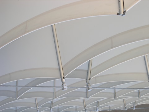 Curved steel beams intersect and support a translucent, light-colored canopy, creating a pattern of repetitive arches. The structure appears modern and industrial, with a minimalist aesthetic that emphasizes geometry and symmetry.