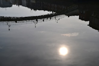 A reflection of a bridge is visible on a body of calm water with the sun casting a bright spot on the surface. Buildings are also reflected in the water, creating an upside-down view. The light and shadow contrast creates a serene atmosphere.