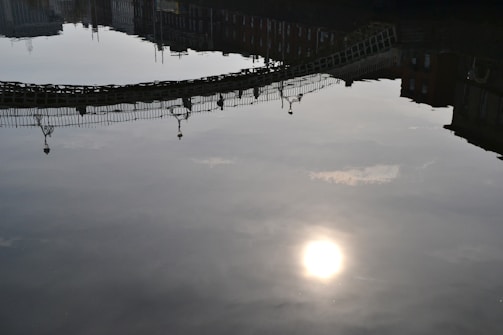 A reflection of a bridge is visible on a body of calm water with the sun casting a bright spot on the surface. Buildings are also reflected in the water, creating an upside-down view. The light and shadow contrast creates a serene atmosphere.