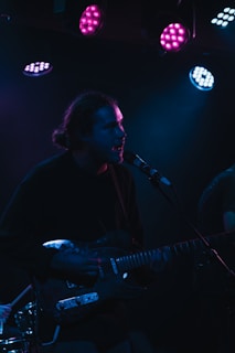 A musician performing on stage, holding an electric guitar and singing into a microphone. The scene is dimly lit with vibrant purple and blue stage lights, creating a moody atmosphere.