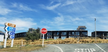 A modern building with multiple windows sits behind a grassy area under a clear blue sky. In the foreground, road signs provide directions to nearby attractions such as hot springs, an ice rink, and holiday accommodations. A stop sign and parking symbol are visible near the road.