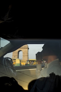 A person is sitting inside a car, seen from a side view, with the silhouette of India Gate visible through the window. There are several yellow and green auto rickshaws in the background, indicating a busy street scene. The person has their hand raised to their face, partially obscuring it.