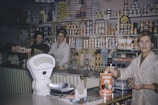 Inside a vintage grocery store, three women are behind a counter filled with various food items. Shelves in the background are stocked with an assortment of canned goods, boxes, and packages. A large, retro weighing scale is visible in the foreground, and one of the women is holding a can labeled 'HACKS'.