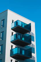 A modern building exterior featuring multiple glass balconies with a sleek design. The structure appears well-maintained and is set against a clear blue sky, suggesting a sunny day. The minimalist architecture emphasizes clean lines and open spaces.