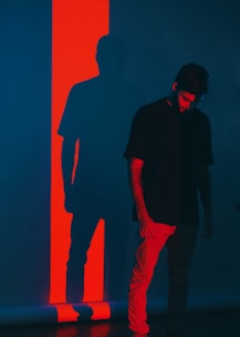 A person stands against a dark background illuminated by a bright red vertical stripe, casting a contrasting shadow. The individual appears to have a contemplative or somber expression, with their head slightly bowed.