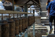 A row of spinning prayer wheels is situated alongside a stone pathway leading up a set of steps. Two people are seen walking up the steps, with their backs to the camera. The background features intricate stone carvings and structures, typical of a religious or cultural site.