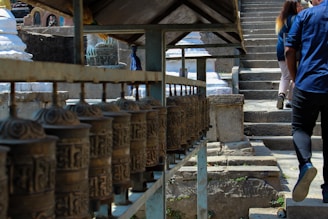 A row of spinning prayer wheels is situated alongside a stone pathway leading up a set of steps. Two people are seen walking up the steps, with their backs to the camera. The background features intricate stone carvings and structures, typical of a religious or cultural site.