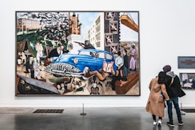 A large, vibrant mural depicting historical and social themes with various figures, including a prominent blue car at the center. Surrounding imagery includes industrial scenes, people in different activities, and symbolic elements that suggest a narrative of struggle and change. Two people stand in front of the painting, possibly discussing or admiring the artwork.