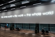 A modern interior space features a large wall with illuminated neon text displaying 'the whole world + the work = the whole world.' Below the text, people are seated on benches, some engaging in conversation, while a person in a blue jacket walks nearby. The floor is wooden, and the overall ambiance suggests a gallery or exhibit environment.