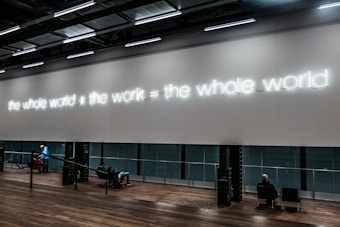 A modern interior space features a large wall with illuminated neon text displaying 'the whole world + the work = the whole world.' Below the text, people are seated on benches, some engaging in conversation, while a person in a blue jacket walks nearby. The floor is wooden, and the overall ambiance suggests a gallery or exhibit environment.