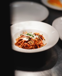 A bowl of spaghetti pasta garnished with sliced cheese and chopped green herbs, served in a white dish. The dish is placed on a dark surface, with portions of other dishes visible in the blurred background.