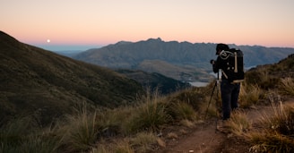 A landscape with a hiker or photographer standing on a dirt trail with a tripod, overlooking a vast valley at dawn or dusk. The scene includes rolling hills and distant mountains under a gradient sky with a visible moon.