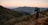 A landscape with a hiker or photographer standing on a dirt trail with a tripod, overlooking a vast valley at dawn or dusk. The scene includes rolling hills and distant mountains under a gradient sky with a visible moon.