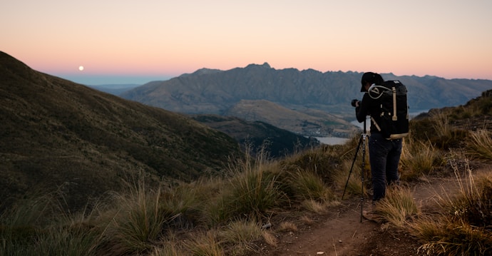 A landscape with a hiker or photographer standing on a dirt trail with a tripod, overlooking a vast valley at dawn or dusk. The scene includes rolling hills and distant mountains under a gradient sky with a visible moon.