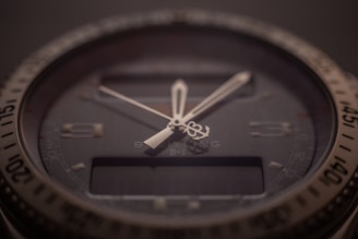 A close-up of a sophisticated wristwatch with a dark dial and luminescent hands. The bezel is marked with numbers, and there is a digital display beneath the hands indicating additional features. The brand name and model are subtly visible on the dial, indicating a premium design.