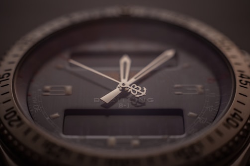 A close-up of a sophisticated wristwatch with a dark dial and luminescent hands. The bezel is marked with numbers, and there is a digital display beneath the hands indicating additional features. The brand name and model are subtly visible on the dial, indicating a premium design.