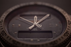 A close-up of a sophisticated wristwatch with a dark dial and luminescent hands. The bezel is marked with numbers, and there is a digital display beneath the hands indicating additional features. The brand name and model are subtly visible on the dial, indicating a premium design.