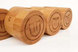 Several cylindrical wooden containers with a smooth, polished finish are aligned in a row. Each container features a circular, engraved logo on top, consisting of stylized geometric shapes. The wood grain is visible, giving a natural and earthy appearance.