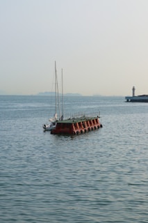 A floating platform anchored in a calm sea with a sailboat nearby. In the distance, a lighthouse is situated on a jetty, and faint outlines of distant hills are visible under a clear sky.