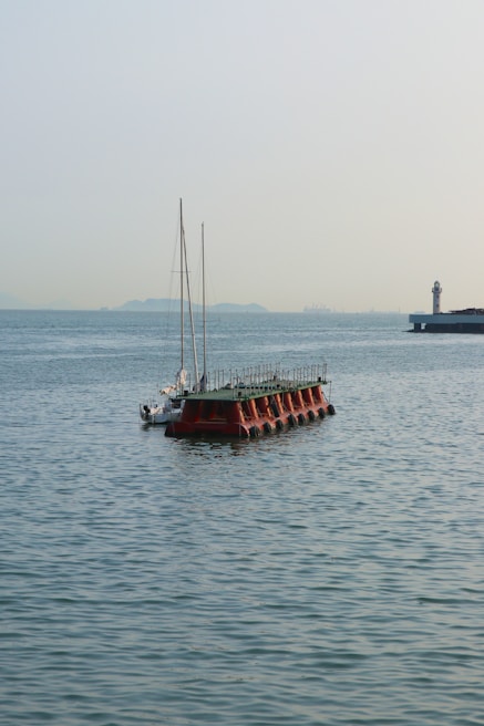 A floating platform anchored in a calm sea with a sailboat nearby. In the distance, a lighthouse is situated on a jetty, and faint outlines of distant hills are visible under a clear sky.