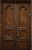 An ornately carved wooden door with geometric designs and a brass handle sits in a rustic frame. The wood is aged, giving it a deep, rich brown color with intricate patterns on both panels.