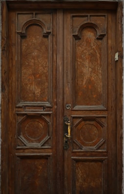 An ornately carved wooden door with geometric designs and a brass handle sits in a rustic frame. The wood is aged, giving it a deep, rich brown color with intricate patterns on both panels.