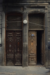 Two ornate, weathered wooden doors side by side with intricate carvings and aged textures. One door is slightly ajar, revealing darkness behind it. The building's facade is covered with an uneven, worn surface, and a blue street number '51' is attached above one door. A round white light or object is centered above the doors.