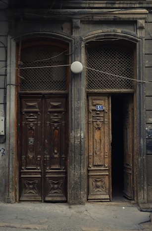 Two ornate, weathered wooden doors side by side with intricate carvings and aged textures. One door is slightly ajar, revealing darkness behind it. The building's facade is covered with an uneven, worn surface, and a blue street number '51' is attached above one door. A round white light or object is centered above the doors.