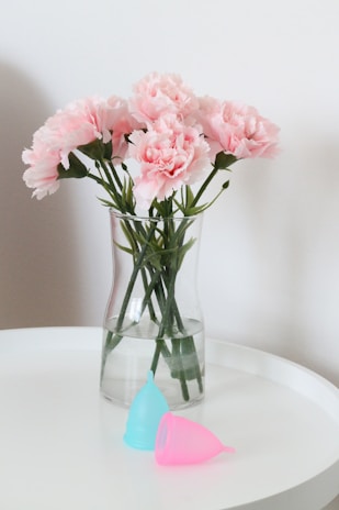 A vase with pink carnations sits on a white round table. Next to the vase are two menstrual cups, one blue and one pink. The background is a plain white wall.