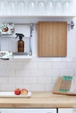 A tidy kitchen area with white tiled walls and a wooden countertop. There is a wooden cutting board hanging on the wall, a brown bottle on a metal rack, dish towels with a sloth pattern, and rows of inverted glass tumblers on a shelf above. On the countertop, there are two apples in a white dish, beeswax wraps, and a beige kitchen cloth.