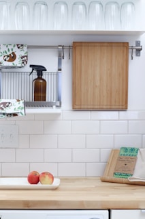 A tidy kitchen area with white tiled walls and a wooden countertop. There is a wooden cutting board hanging on the wall, a brown bottle on a metal rack, dish towels with a sloth pattern, and rows of inverted glass tumblers on a shelf above. On the countertop, there are two apples in a white dish, beeswax wraps, and a beige kitchen cloth.