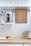 A tidy kitchen area with white tiled walls and a wooden countertop. There is a wooden cutting board hanging on the wall, a brown bottle on a metal rack, dish towels with a sloth pattern, and rows of inverted glass tumblers on a shelf above. On the countertop, there are two apples in a white dish, beeswax wraps, and a beige kitchen cloth.