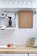 A tidy kitchen area with white tiled walls and a wooden countertop. There is a wooden cutting board hanging on the wall, a brown bottle on a metal rack, dish towels with a sloth pattern, and rows of inverted glass tumblers on a shelf above. On the countertop, there are two apples in a white dish, beeswax wraps, and a beige kitchen cloth.