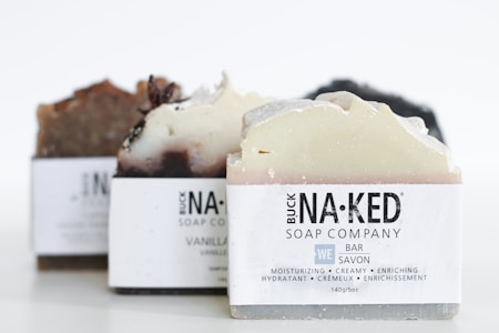 Several bars of artisanal soap with varying shades of brown, cream, and white are neatly arranged. Each soap bar is labeled with 'Buck Naked Soap Company' and descriptions like moisturizing, creamy, and enriching are visible.