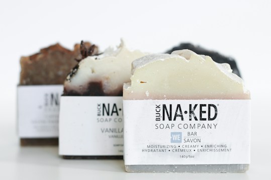 Several bars of artisanal soap with varying shades of brown, cream, and white are neatly arranged. Each soap bar is labeled with 'Buck Naked Soap Company' and descriptions like moisturizing, creamy, and enriching are visible.