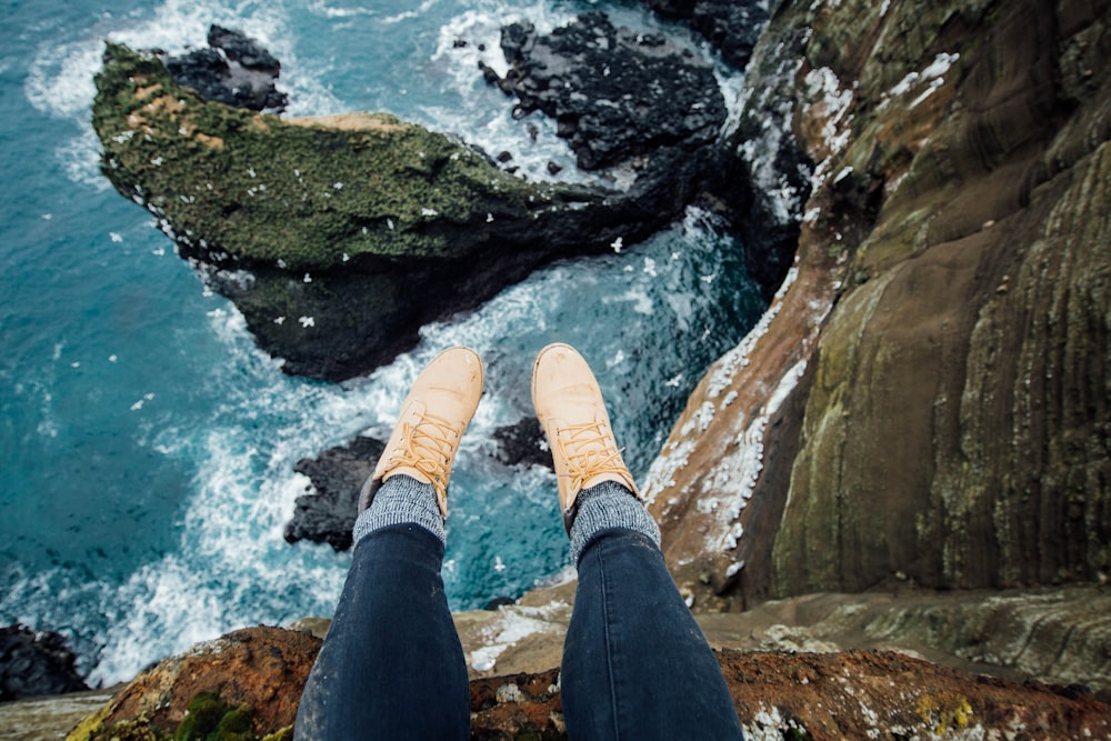 person wearing blue denim jeans sitting on rocky hill viewing sea