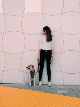 A person wearing a white top and black pants stands against a wall with a geometric pattern. They are holding a red leash attached to a small, fluffy dog wearing a colorful harness. The floor features a bright yellow tile pattern.