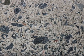 A close-up texture of a concrete surface with various stone aggregates embedded. The stones vary in size and color, predominantly in shades of black, gray, and some brownish tones. The texture is rough with visible specks and patches, giving it a natural and rugged appearance.