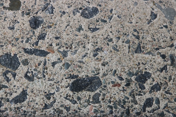 A close-up texture of a concrete surface with various stone aggregates embedded. The stones vary in size and color, predominantly in shades of black, gray, and some brownish tones. The texture is rough with visible specks and patches, giving it a natural and rugged appearance.