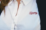 A person wearing a white collared shirt with the embroidered name 'Tommy' in red on the chest. The shirt has buttons and there is a hint of brown hair visible on the left side.