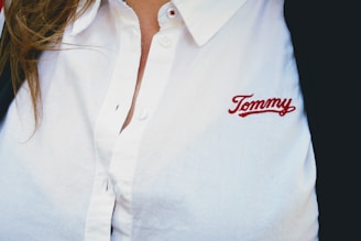 A person wearing a white collared shirt with the embroidered name 'Tommy' in red on the chest. The shirt has buttons and there is a hint of brown hair visible on the left side.