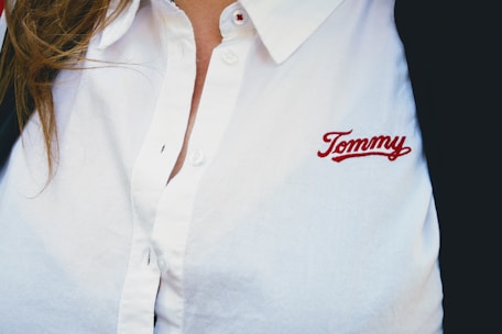 A person wearing a white collared shirt with the embroidered name 'Tommy' in red on the chest. The shirt has buttons and there is a hint of brown hair visible on the left side.