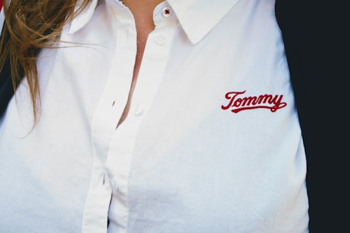 A person wearing a white collared shirt with the embroidered name 'Tommy' in red on the chest. The shirt has buttons and there is a hint of brown hair visible on the left side.