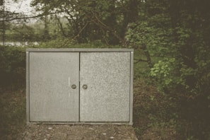 A sleek metal outdoor cabinet storage standing tall against a wooden fence with garden accessories