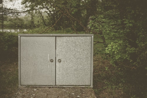 A metallic utility cabinet stands in a natural setting, surrounded by lush green foliage. The surface of the cabinet is weathered, with a textured, slightly rusty appearance. It is placed on a small paved area, indicating an outdoor installation, likely for electrical or telecommunications purposes.