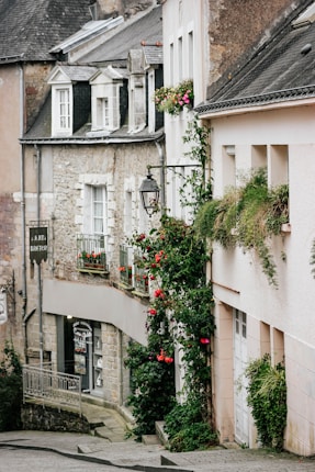 A picturesque narrow street lined with charming old buildings, featuring stone and plaster facades and decorative window planters filled with vibrant flowers. Ivy and climbing plants embellish the walls, while vintage-style lanterns add a touch of classic elegance.