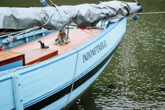 A docked sailboat with 'INDOMPTABLE' written on the side appears secured and moored with various ropes. The deck is wooden with some visible objects, and the hull is painted light blue. The water around the boat is calm, appearing greenish in color.